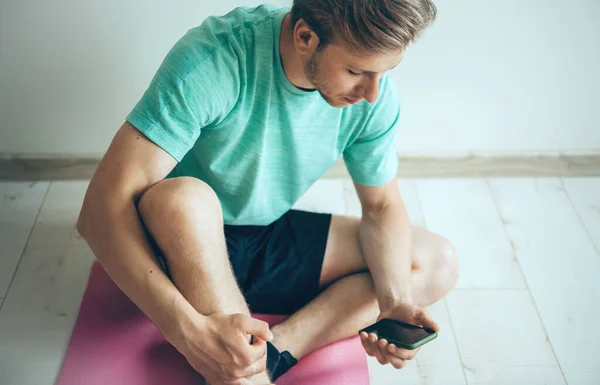 Bionda caucasico uomo indossa abbigliamento sportivo sul pavimento è rilassante mentre si fa fitness a casa e utilizzando un telefono — Foto Stock