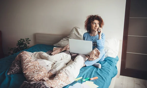 Mujer joven con el pelo rizado está hablando por teléfono y el uso de un ordenador portátil en la cama temprano en la mañana con un pijama azul y haciendo la tarea — Foto de Stock