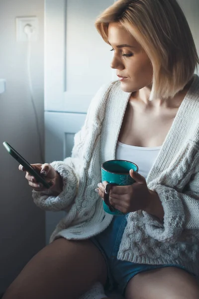 De cerca la foto de una rubia encantadora dama en jersey de punto sentado en la cocina y bebiendo un té mientras charlaba por teléfono —  Fotos de Stock