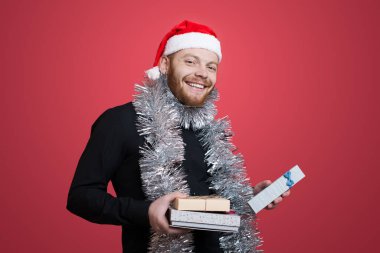 Kızıl saçlı, sakallı, noel kıyafetleri ve süslemeleri olan beyaz adam kırmızı bir stüdyo duvarında hediyeler tutarken kameraya gülümsüyor.