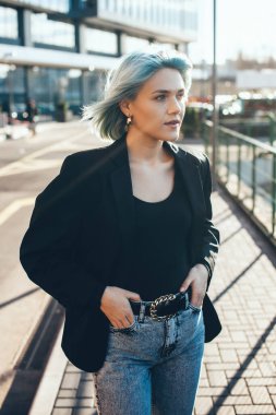 Mavi saçlı kadın, şehrin dışında, modern bir binanın yanında güneş ışığında poz veriyor.