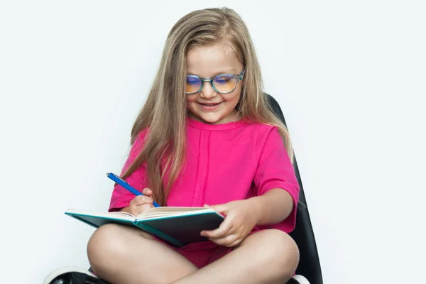 Chica rubia caucásica con anteojos escribiendo algo en un libro mientras usa camisa roja en una pared de estudio blanca y sentado en un sillón — Foto de Stock