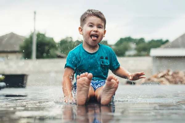 Foto vista frontal de um menino caucasiano gesticulando felicidade com a língua enquanto brincava descalço na água no chão após a chuva — Fotografia de Stock