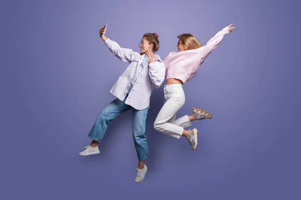 Zwei charmante Frauen in heller Kleidung und blonden Haaren springen auf eine violette Studiowand und machen ein Selfie mit dem Handy — Stockfoto