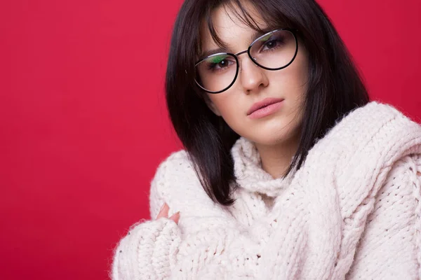 De cerca foto de una mujer morena caucásica con gafas posando en un suéter caliente en una pared de estudio rojo abrazándose —  Fotos de Stock