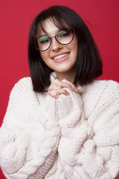 Entzückende brünette Frau mit Brille in einem gestrickten weißen Pullover posiert auf einer roten Studiowand, die Hände zusammen lächelnd in die Kamera — Stockfoto