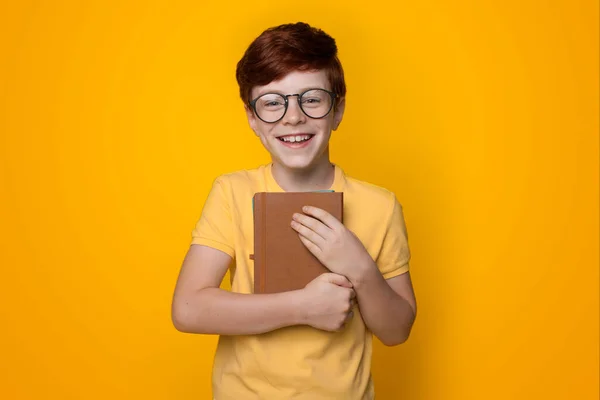 Close up foto de um menino gengibre abraçando livro e usar óculos sorrindo para a câmera em uma parede de estúdio amarelo — Fotografia de Stock