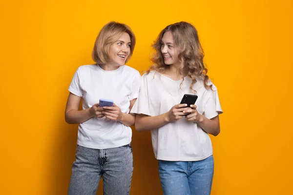 Glada blonda damer chattar på telefonen och ler mot varandra på en gul ateljé vägg i vita t-shirts — Stockfoto