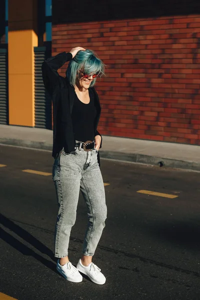 Jonge blauwe harige vrouw wandelen in de straat met bril en glimlach met een bakstenen muur op de achtergrond in een zonnige dag — Stockfoto