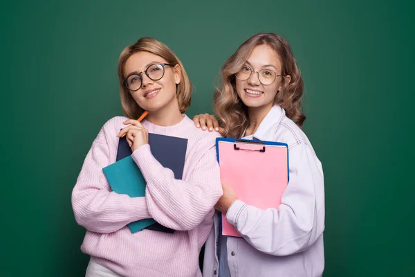 Närbild foto av två blonda systrar med glasögon som ler mot kameran poserar på en grön studiovägg med några skolmappar — Stockfoto