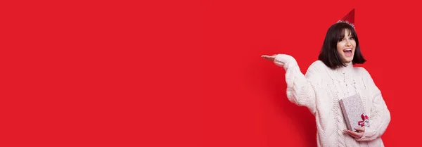 Brunett dam reklam något som håller i handflatan på en röd studio vägg med en gåva i handen och fest hatt nära ledigt utrymme — Stockfoto