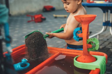 Tişörtsüz beyaz bir çocuk bahçesinde suyla oynuyor. Oyuncak karga kullanarak bir kutuya toprak ekliyor.