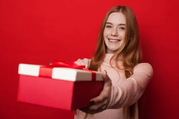 Mujer caucásica con pecas y cabello de jengibre está dando una caja con regalo en la cámara y sonrisa en la pared del estudio rojo — Foto de Stock