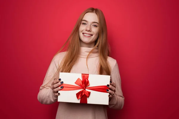 Hermosa mujer caucásica con el pelo rojo y pecas sonriendo a la cámara y sosteniendo un regalo en las manos posando en una pared de estudio rojo — Foto de Stock