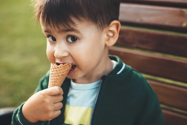 Close up foto de um menino morena comendo um sorvete enquanto sentado no banco no parque e olhando para a câmera — Fotografia de Stock