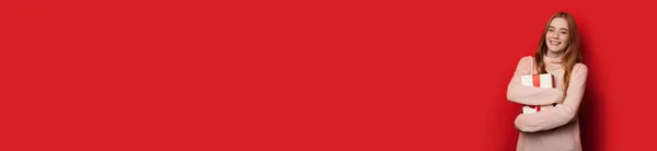 Nettes Foto einer Ingwerfrau mit Sommersprossen, die ein Geschenk auf einer roten Studiowand mit freiem Platz umarmt — Stockfoto