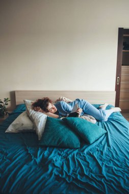 Mavi pijamalı kıvırcık saçlı kadın yatakta kitap okuyor. Hafta sonunun tadını çıkarıyor.