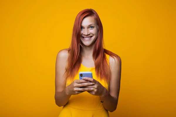 Mujer jengibre feliz sonriendo a la cámara en un vestido amarillo sosteniendo un teléfono y charlando en él en una pared de estudio — Foto de Stock
