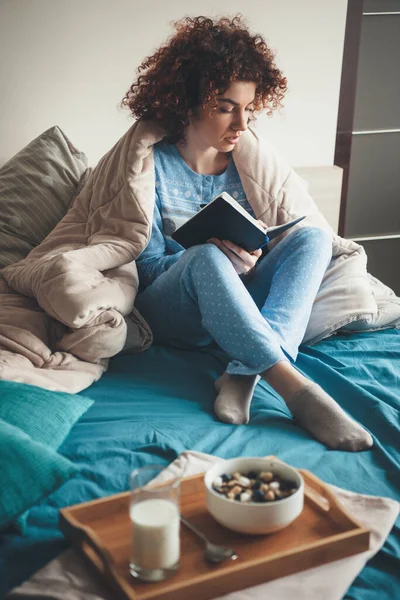 Mujer caucásica con el pelo rizado está leyendo un libro en la cama con un pijama azul cubierto con una colcha antes de comer cereales con leche — Foto de Stock