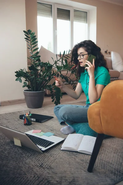 Lovely curly haired woman talking on phone while sitting on floor and doing homework with a computer and some books — Stock Photo, Image