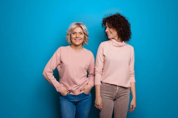 Zwei kaukasische Frauen mit blonden und brünetten lockigen Haaren tragen dieselben Pullover an einer blauen Studiowand und lächeln in die Kamera — Stockfoto