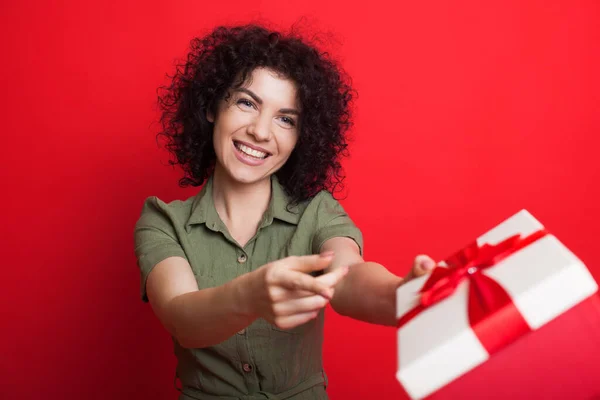 Encantadora mujer de pelo rizado sonriendo y dando un regalo a la cámara haciendo gestos con el dedo que es para usted mientras posa en una pared de estudio rojo — Foto de Stock