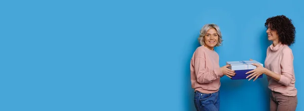 stock image Caucasian blonde lady giving a present to her brunette curly haired friend posing on a blue wall with free space advertising something