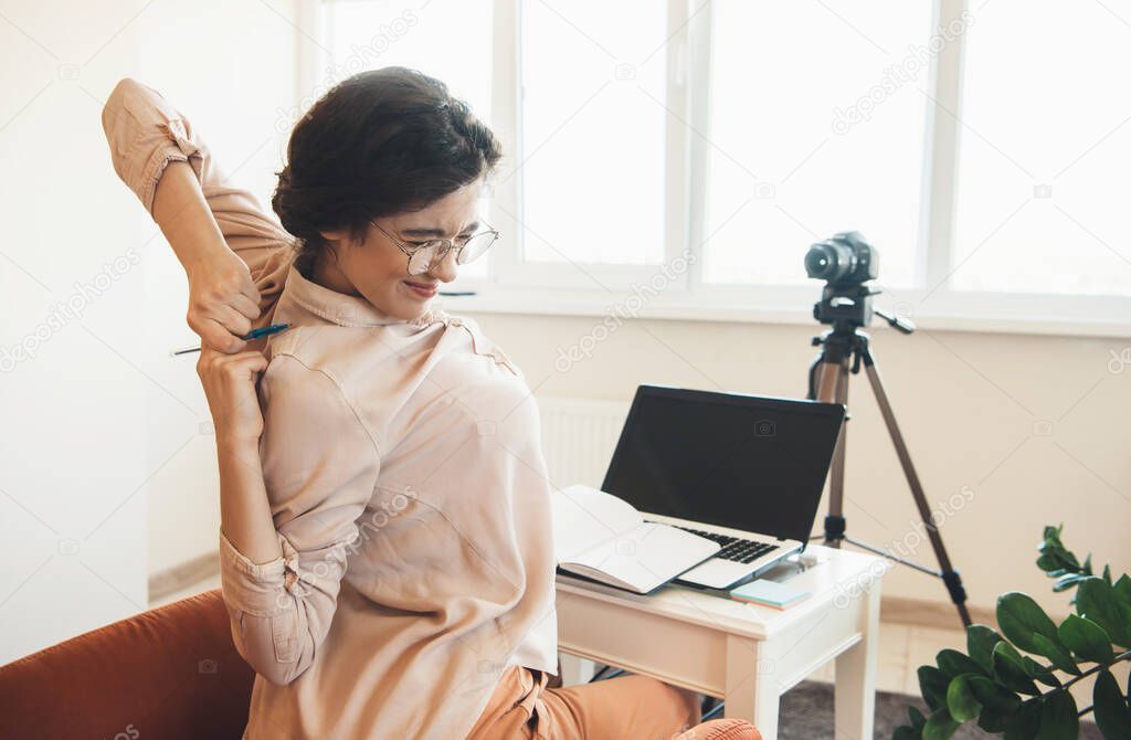 Tired caucasian woman with eyeglasses having online lessons using a camera and laptop while stretching