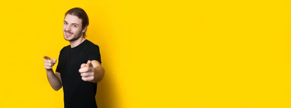 Homem caucasiano barbudo em uma camiseta preta apontando para a câmera e sorrir em um banner de estúdio amarelo — Fotografia de Stock