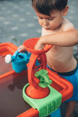 Beyaz tenli çıplak çocuk bahçede plastik bir oyuncak ve suyla oynuyor.