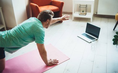 Dijital fitness seansından önce yerde dizüstü bilgisayar ve bir bardak su kullanarak ısınan beyaz bir adamın üst manzara fotoğrafı.