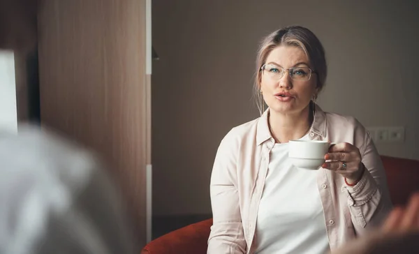 Señora caucásica mayor bebiendo una taza de café y discutiendo con alguien usando anteojos y ropa casual en casa — Foto de Stock