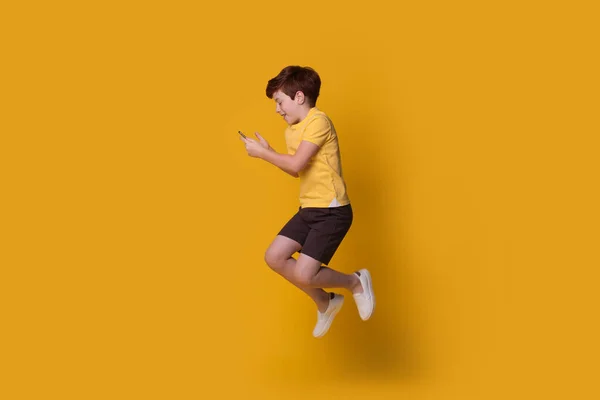 Gengibre caucasiano menino vestindo uma camiseta amarela está pulando na parede do estúdio enquanto conversa no celular — Fotografia de Stock