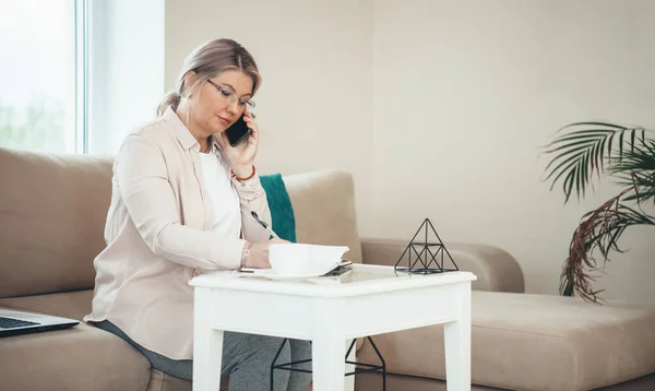 Side view photo of a senior businesswoman talking on phone while writing something and sitting on the couch near the laptop — Stock Photo, Image