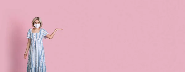 Jovem caucasiana com cabelo loiro e máscara médica no rosto está segurando algo na palma da mão posando em um banner rosa no estúdio — Fotografia de Stock