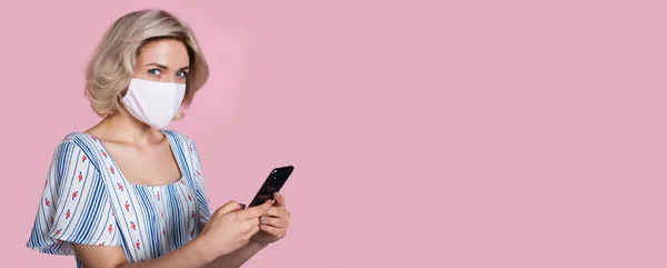 Blonde woman advertising something while posing on a pink studio wall with free space and wear mask on face and holding a phone