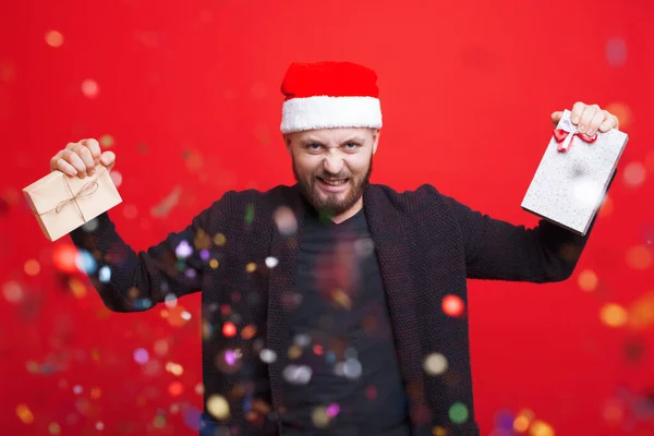 Érzelmi kaukázusi férfi szakállal és télapósapkával két ajándékot tart a vörös falon konfetti gesztusokkal. — Stock Fotó