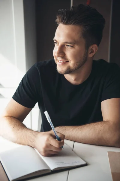 Sonriente freelancer caucásico escribiendo algo en el libro mientras piensa y mira a través de la ventana en casa —  Fotos de Stock