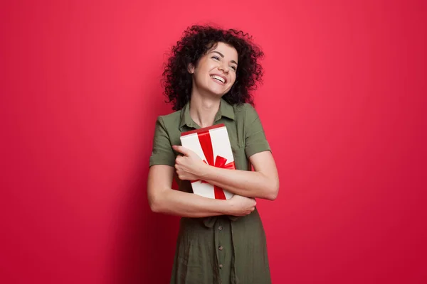 Mujer linda en un vestido y cabello rizado abrazando un regalo y sonrisa en una pared roja en el estudio — Foto de Stock