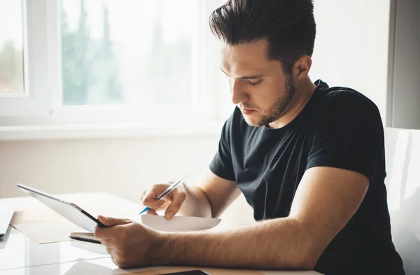 Skäggig kaukasisk affärsman som arbetar hemifrån med en surfplatta och några papper i svart t-shirt i vardagsrummet — Stockfoto