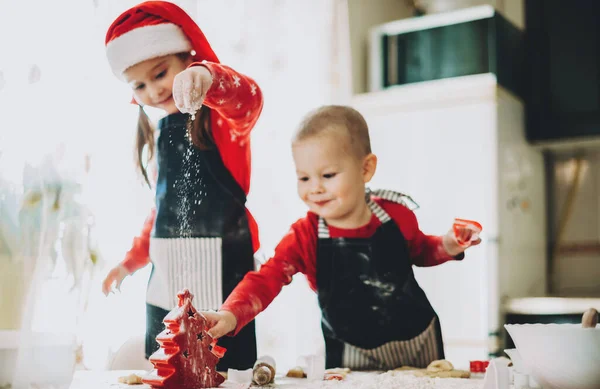 Preparación navideña de dos hermanos haciendo galletas vestidas con ropas rojas con sombrero de santa — Foto de Stock