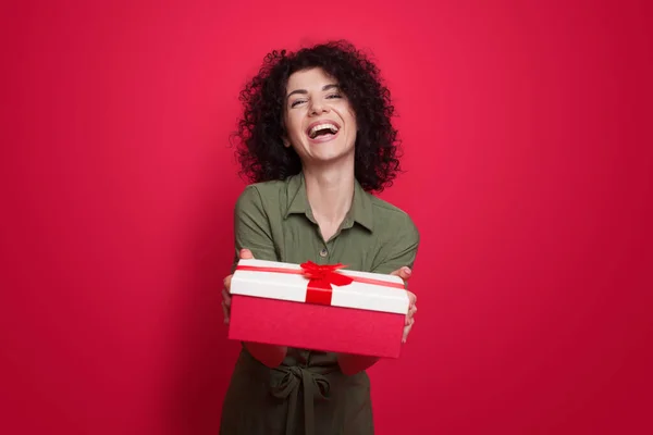 Encantadora mujer morena con el pelo rizado dando un regalo a la cámara sonriendo en una pared de estudio rojo — Foto de Stock