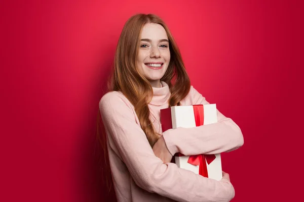 Linda mujer de jengibre con pecas abrazando un regalo y sonrisa en la cámara en la pared del estudio rojo — Foto de Stock