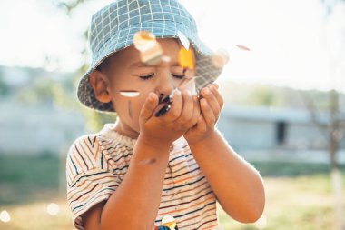 Kafkasyalı çocuk güneşli bir günde dışarıda şapka takarken konfeti üflüyor.