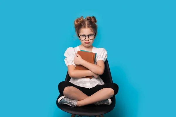 Funny caucásico escuela chica gesto decepción en un estudio azul de la pared, mientras que el uso de gafas y sentado en sillón celebración de libros —  Fotos de Stock