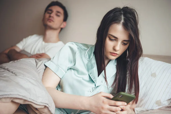 Caucásico hombre cuidando de su esposa mensajes de texto de un mensaje en el teléfono mientras está acostado en la cama mostrando celos — Foto de Stock