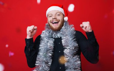 Noel Baba şapkalı sakallı beyaz adam kırmızı stüdyo duvarına yeni yıl süslemeleri takarken gülümsüyor ve mutluluk işareti yapıyor.