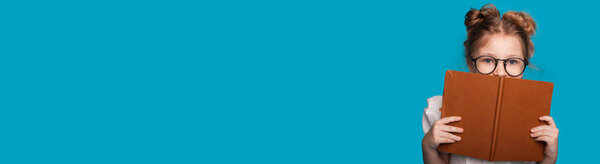 Caucasian girl covering her face with a book while wear eyeglasses is posing on a blue studio wall with free space advertising something