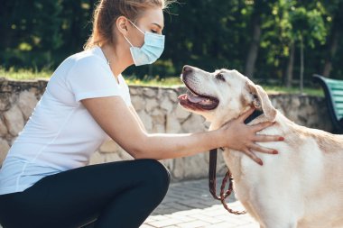 Genç sarışın kadın yüzüne maske takıp parkta yürürken köpeğine dokunuyor.