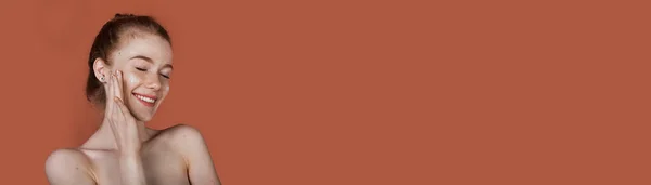 Веснушчатая белая женщина с рыжими волосами и голыми плечами позирует на красной стене студии с свободным пространством, улыбаясь и нанося антивозрастной крем на лицо — стоковое фото